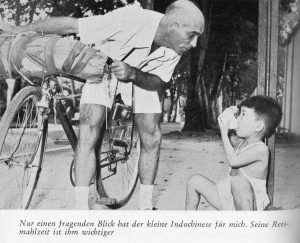 Aufnahme eines schwarz-weiß Foto, bei dem er sich vom Fahrrad zu einem Indochinesischen Jungen runterbeugt
